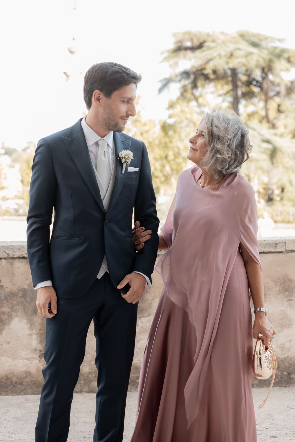 Groom in a navy suit with a white boutonniere stands beside an older woman in a dusty rose dress and shawl at an outdoor wedding.