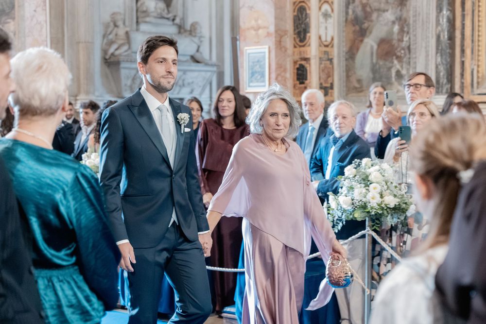 A groom in a navy suit walks hand in hand with an older woman in a blush shawl down the aisle of an ornate church as guests look on.