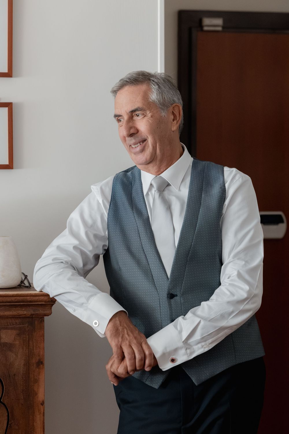 Smiling gray-haired man in a blue-gray vest and white dress shirt leans on a wooden cabinet, looking off to the side in a bright room.