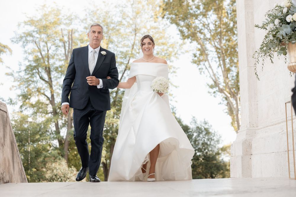 Bride in an elegant off-the-shoulder white gown holding a bouquet walks beside a man in a navy suit outdoors near trees and floral decor.