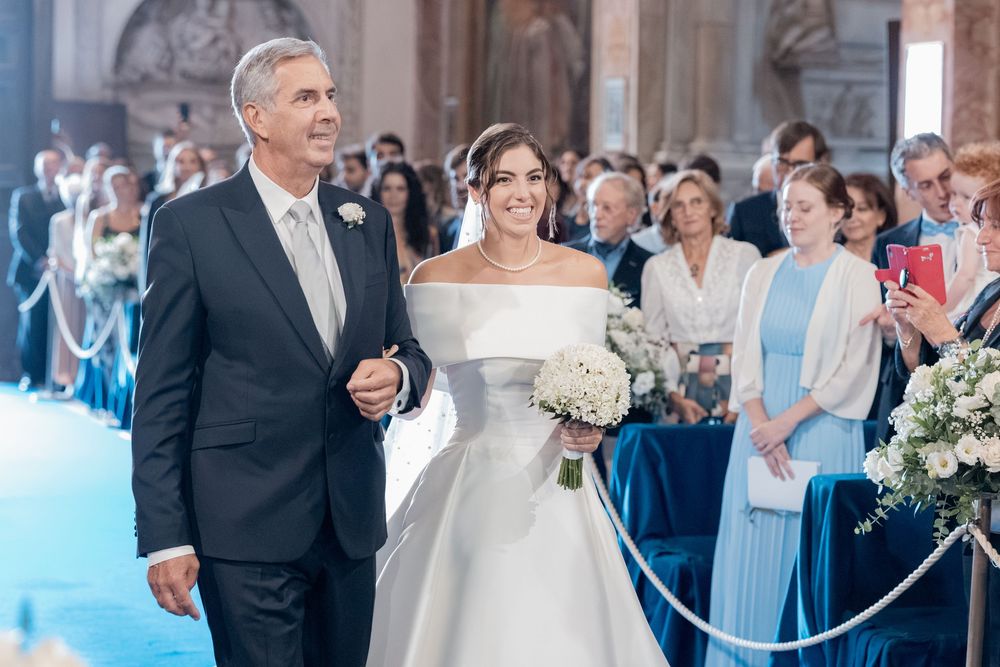Smiling father in a gray suit walks his bride in a white off-shoulder gown and bouquet down the aisle as wedding guests look on.