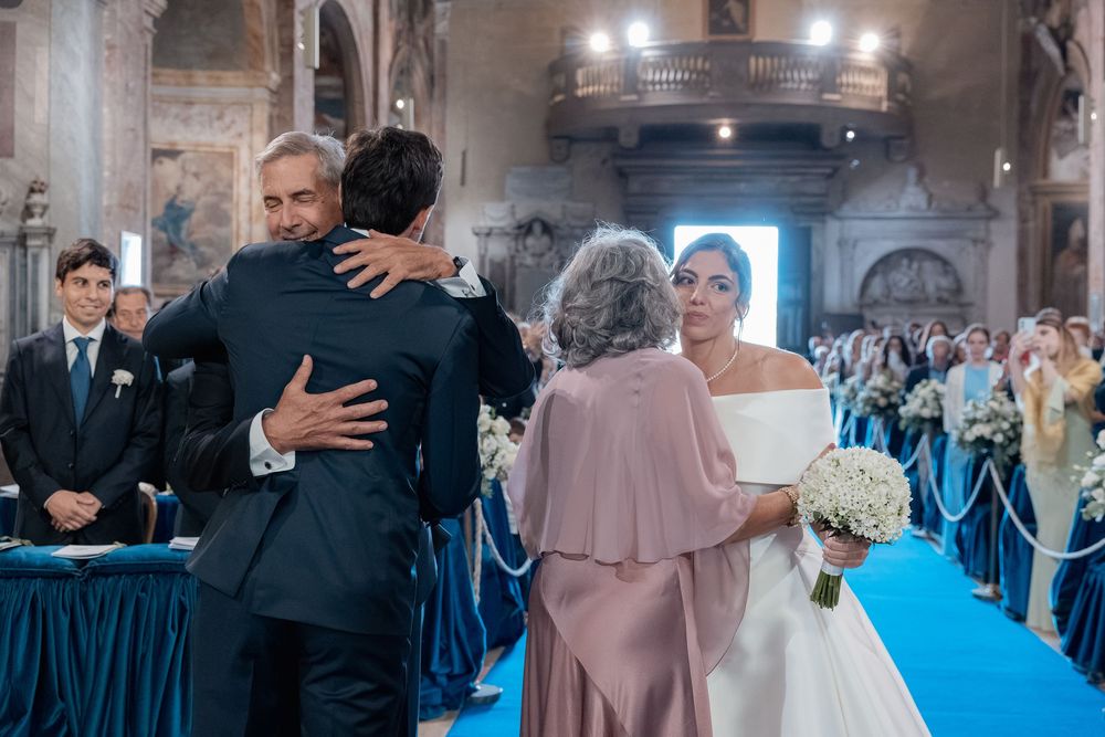 A bride in a white gown holding a bouquet watches as two men embrace in front of a woman in pink during a church wedding ceremony.