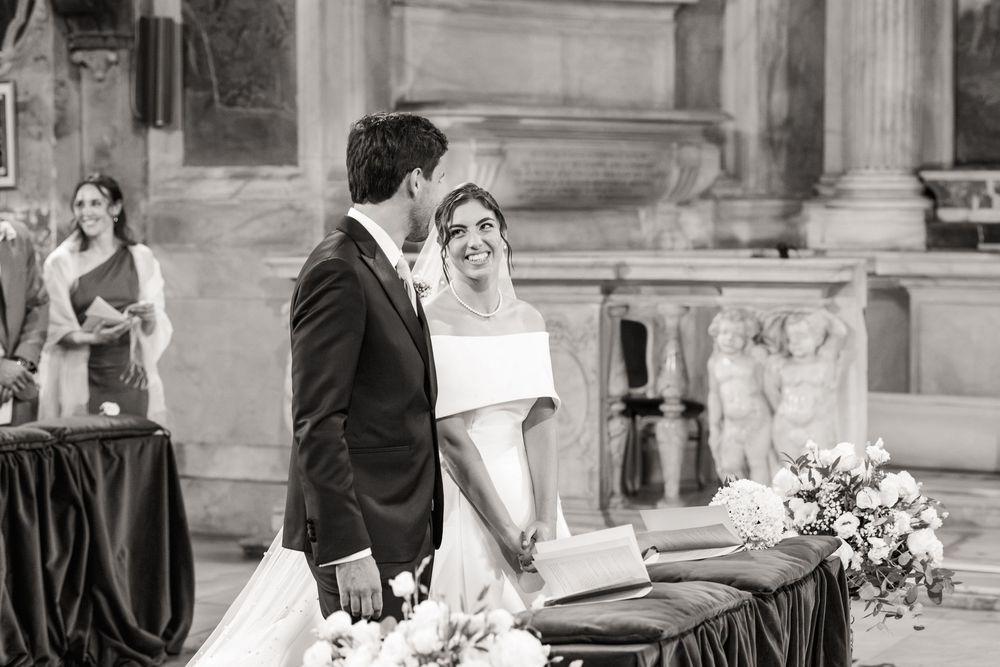 Black and white photo of a bride and groom standing at the altar during a wedding ceremony in a church, with flowers and a guest behind.