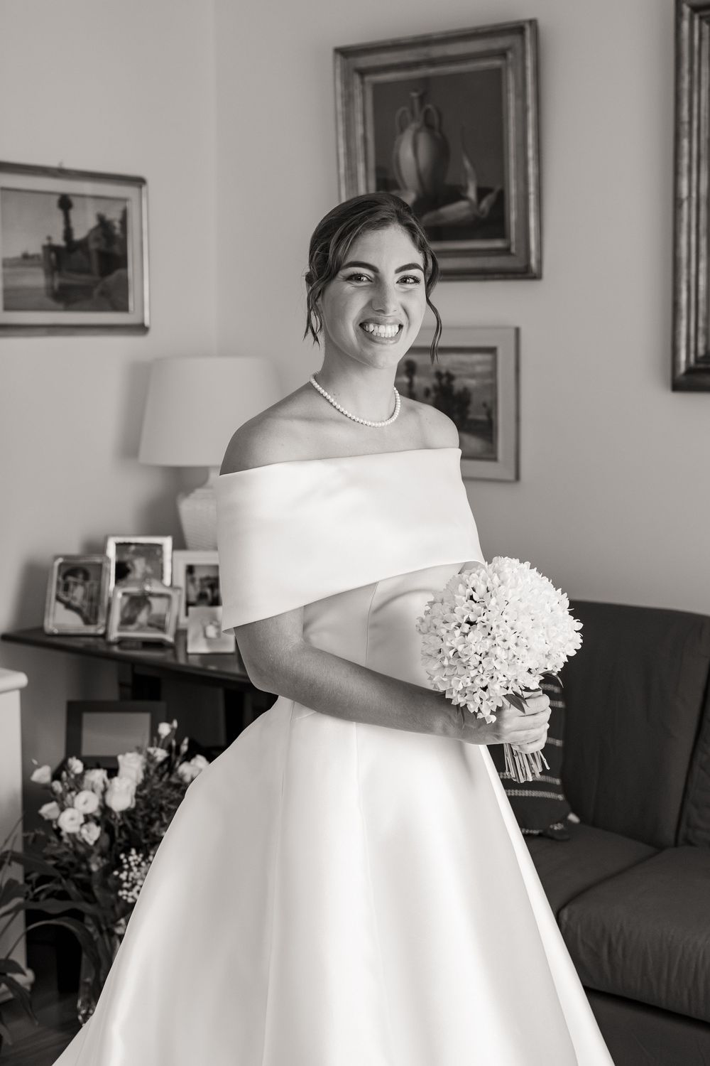 Smiling bride in an off-the-shoulder satin wedding gown and pearl necklace holding a white bouquet in a cozy living room.