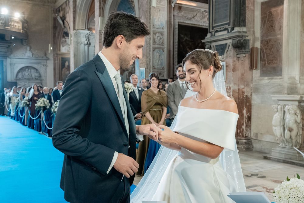 Bride in an off-shoulder white gown and groom in a dark suit exchange rings during a wedding ceremony in an ornate church with guests.