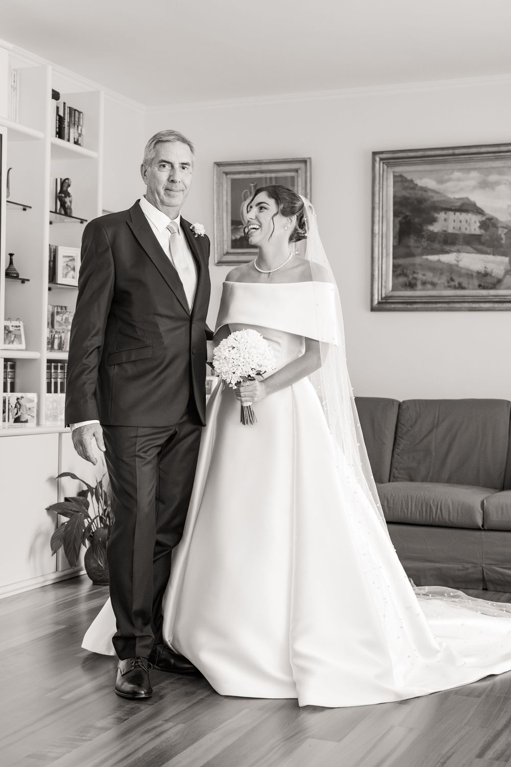 Smiling bride in a strapless white wedding gown and long veil holding a bouquet beside a man in a dark suit in a bright living room.