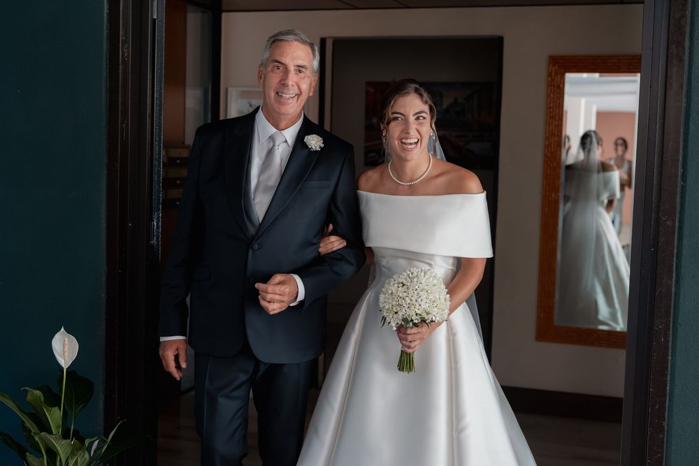 A smiling bride in an off-the-shoulder white wedding gown holds a bouquet while walking arm in arm with a man in a dark suit.