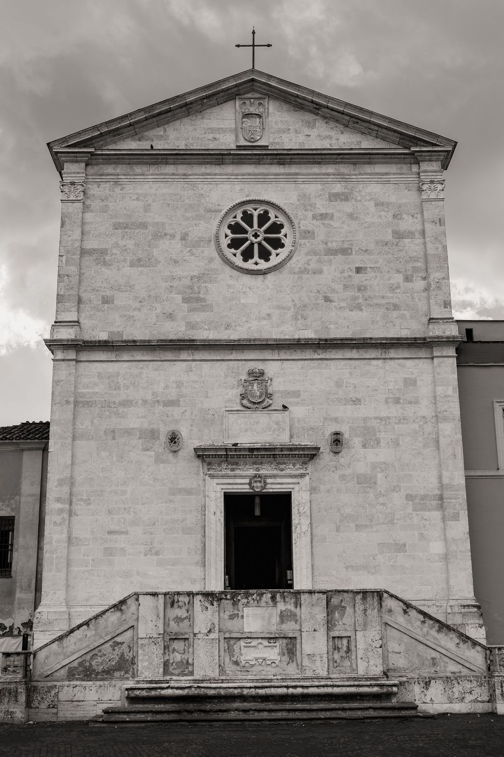 Black and white photo of a historic stone church facade with a rose window, triangular pediment, cross, and a stepped entrance staircase.