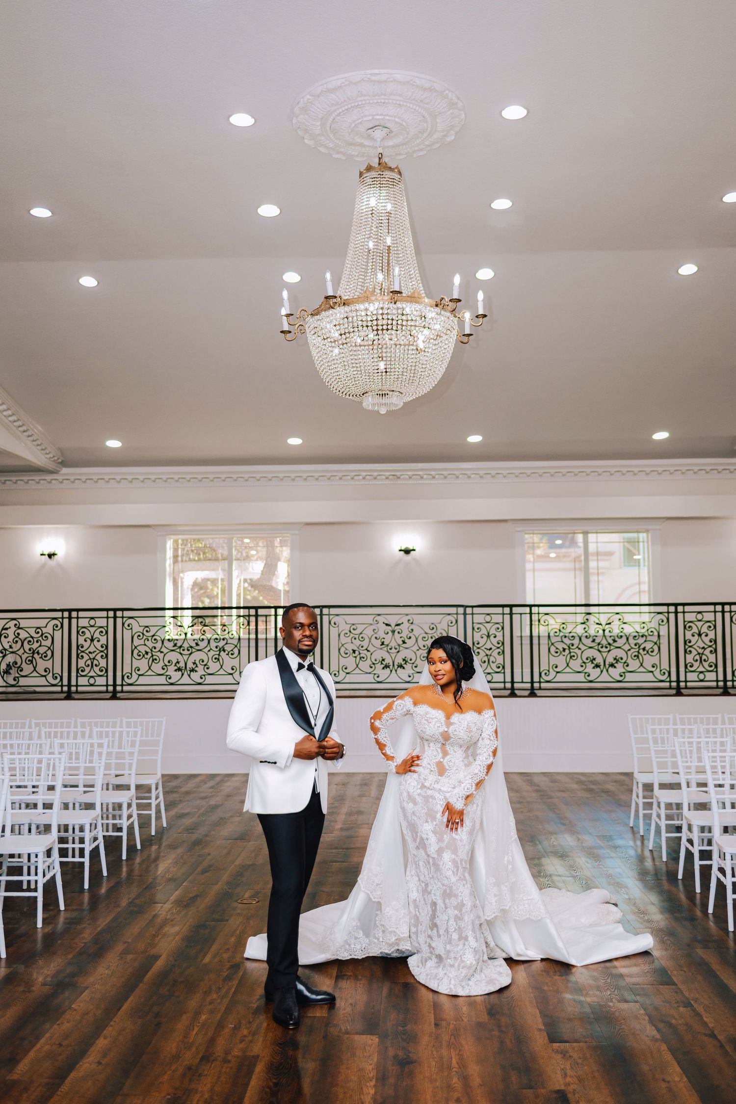 Bride in a lace gown with a long veil and groom in a white tuxedo jacket pose in an elegant banquet hall with a crystal chandelier.