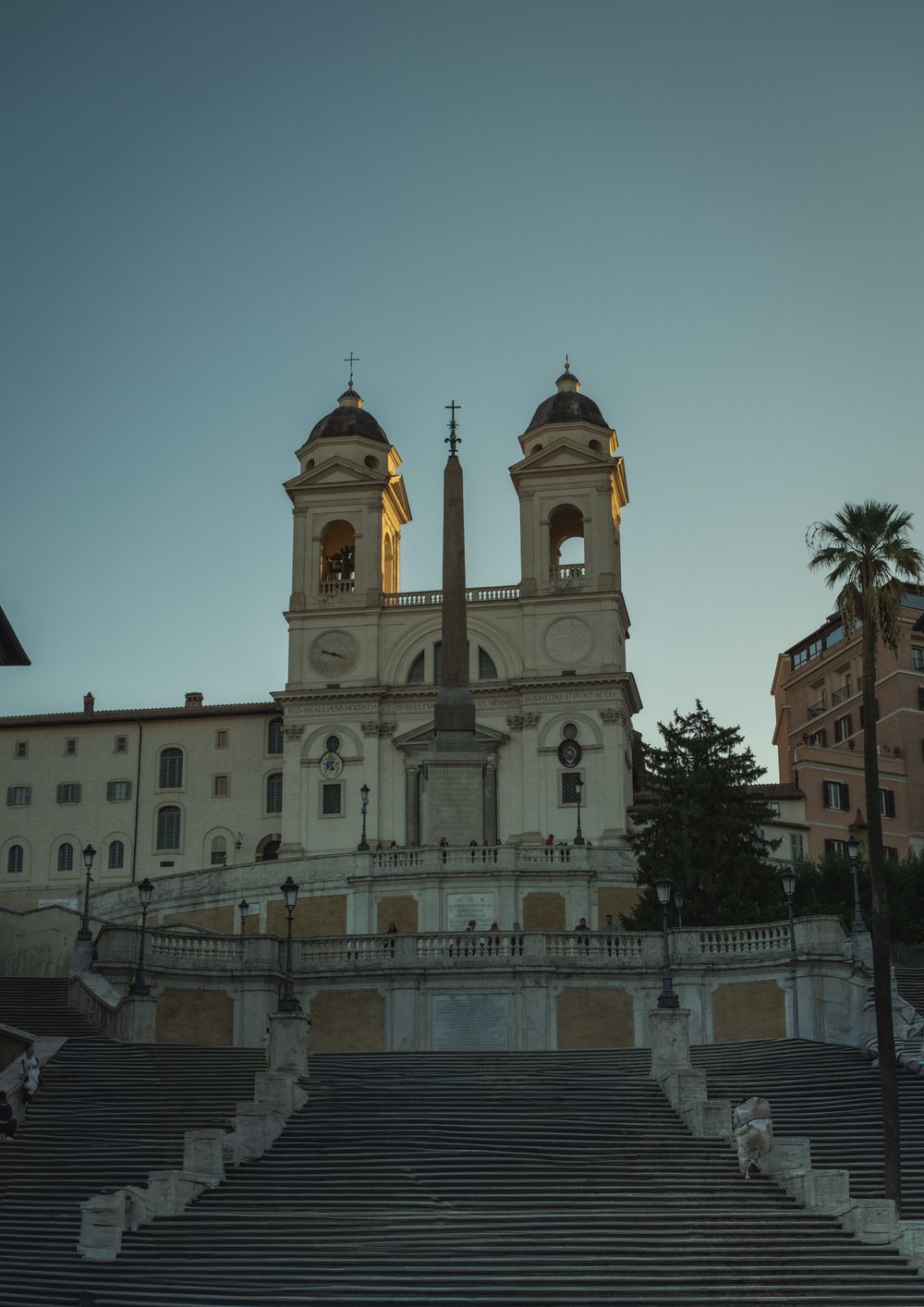 Twin-towered Trinità dei Monti church and obelisk rising above the Spanish Steps in Rome at dusk, with a palm tree beside the stairs.
