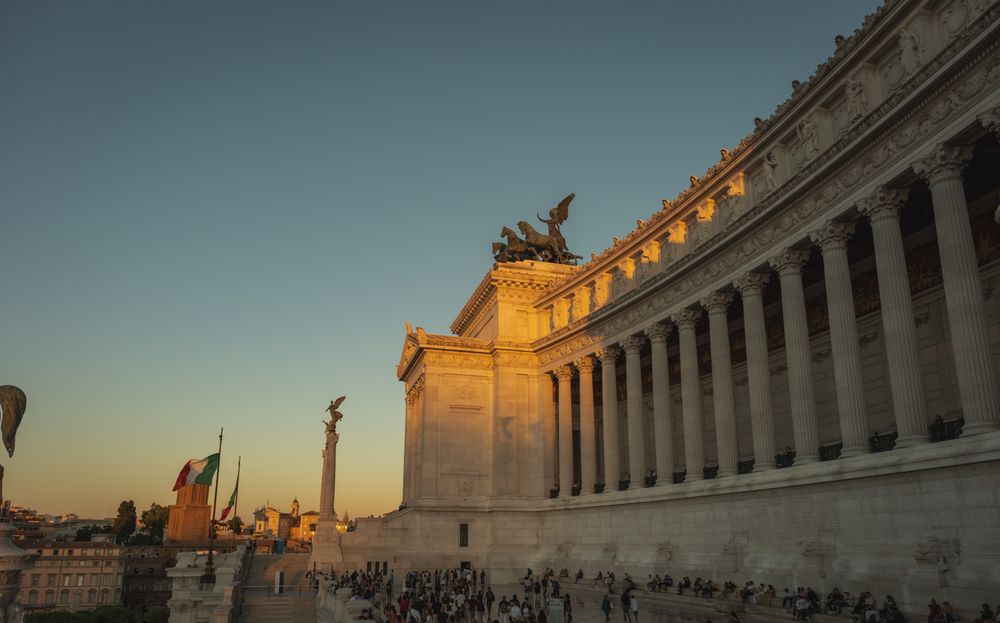 Golden sunset light illuminates the colonnade and bronze quadriga sculpture of the Vittoriano monument in Rome as visitors gather below.