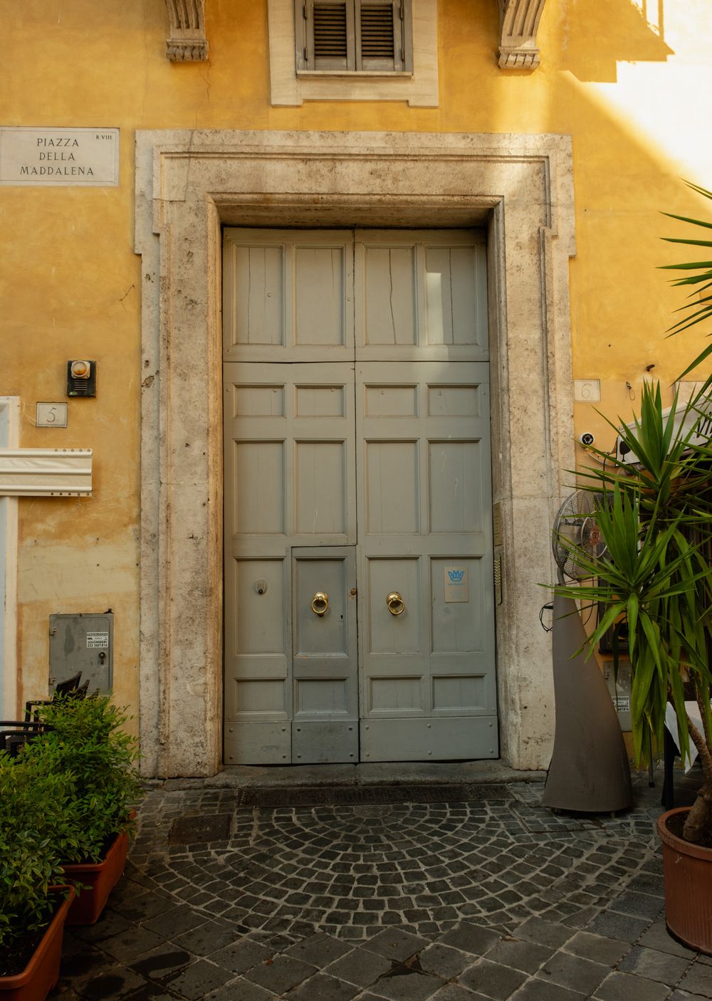Large gray wooden double door set in a stone frame on a yellow building at Piazza della Maddalena, with potted plants and cobblestones.