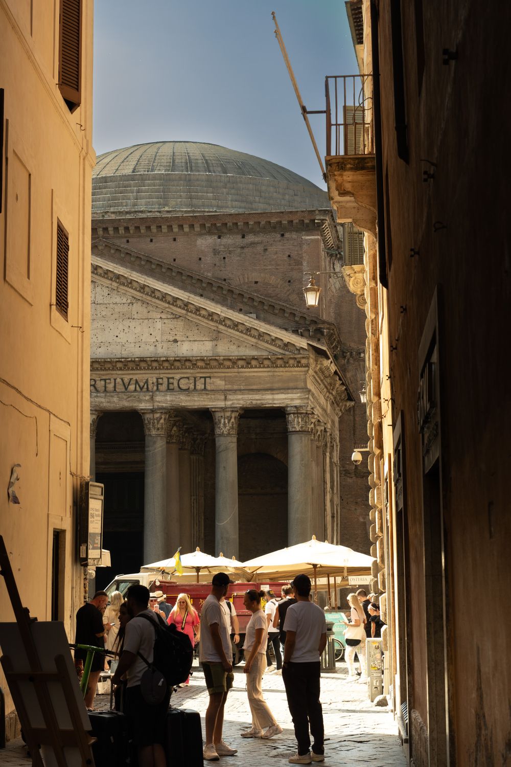 Tourists gather under white umbrellas in a narrow Roman alley framing the columned portico and dome of the Pantheon in Rome, Italy.
