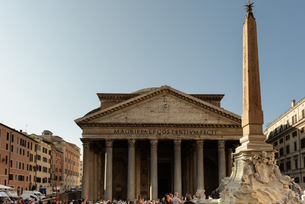 Ancient Roman Pantheon with its columned portico and inscribed pediment, beside an Egyptian obelisk and fountain in a crowded Rome piazza.