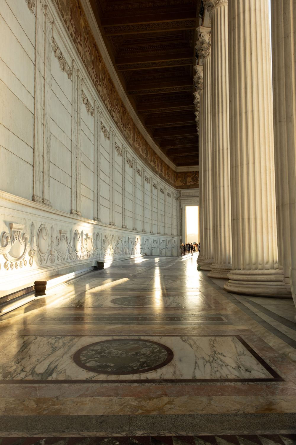 Sunlit marble colonnade with tall fluted columns, ornate carved walls and a polished mosaic floor stretching toward distant visitors.