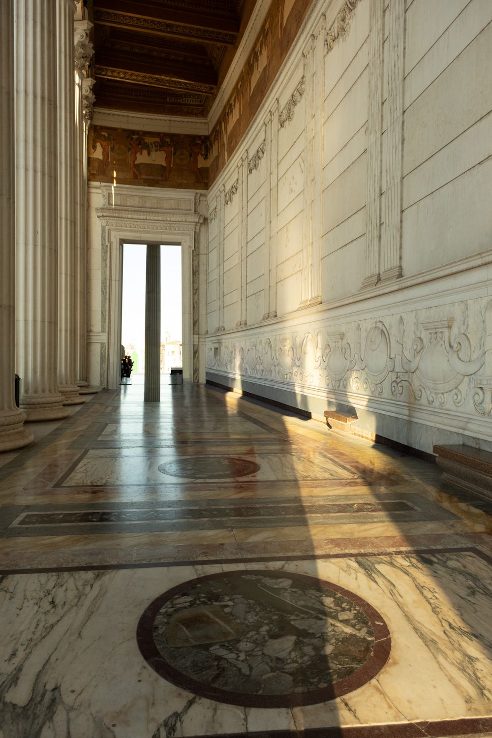 Sunlit marble colonnade corridor with polished inlaid stone floor, carved white relief walls, and a coffered ceiling leading to a bright doo