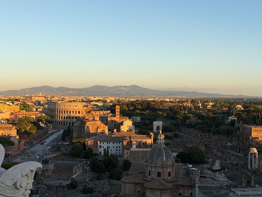 Aerial view of Rome at golden hour showing the Colosseum, ancient Roman Forum ruins, domed churches and distant mountains under a clear sky.