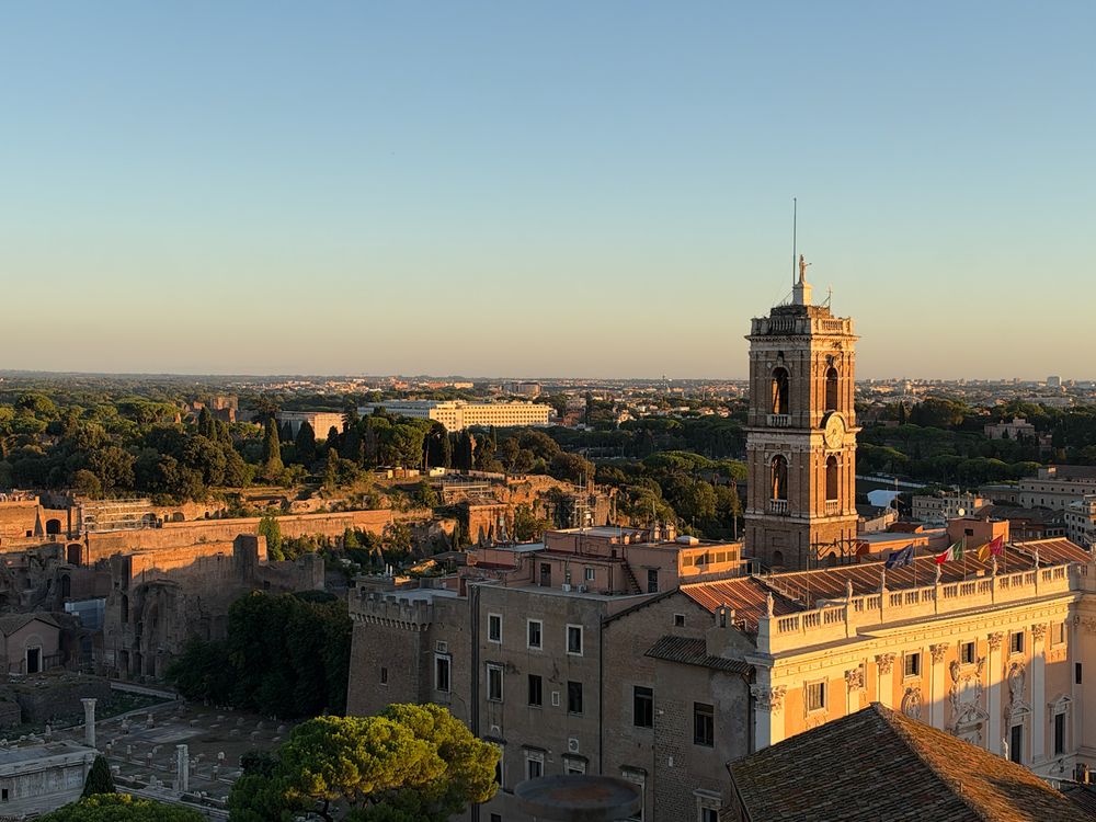 Golden hour view over Rome rooftops with a historic bell tower, ancient ruins and lush green trees stretching toward the distant horizon.