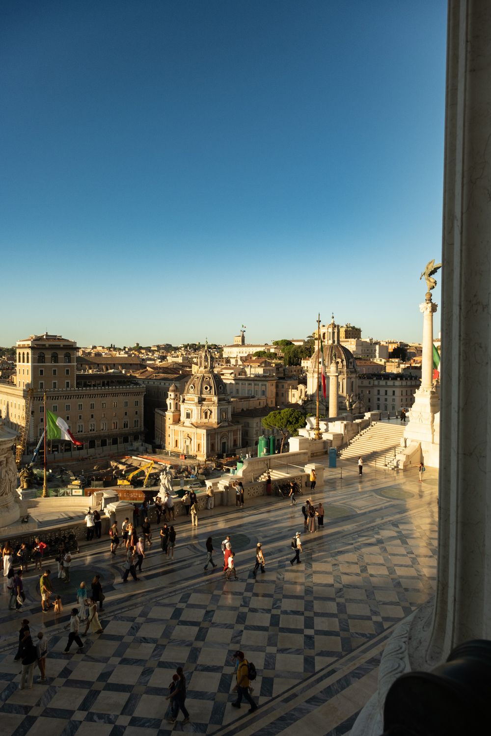 Tourists stroll across a checkered marble terrace overlooking Rome's historic rooftops, domed churches, and a tall column at golden hour.