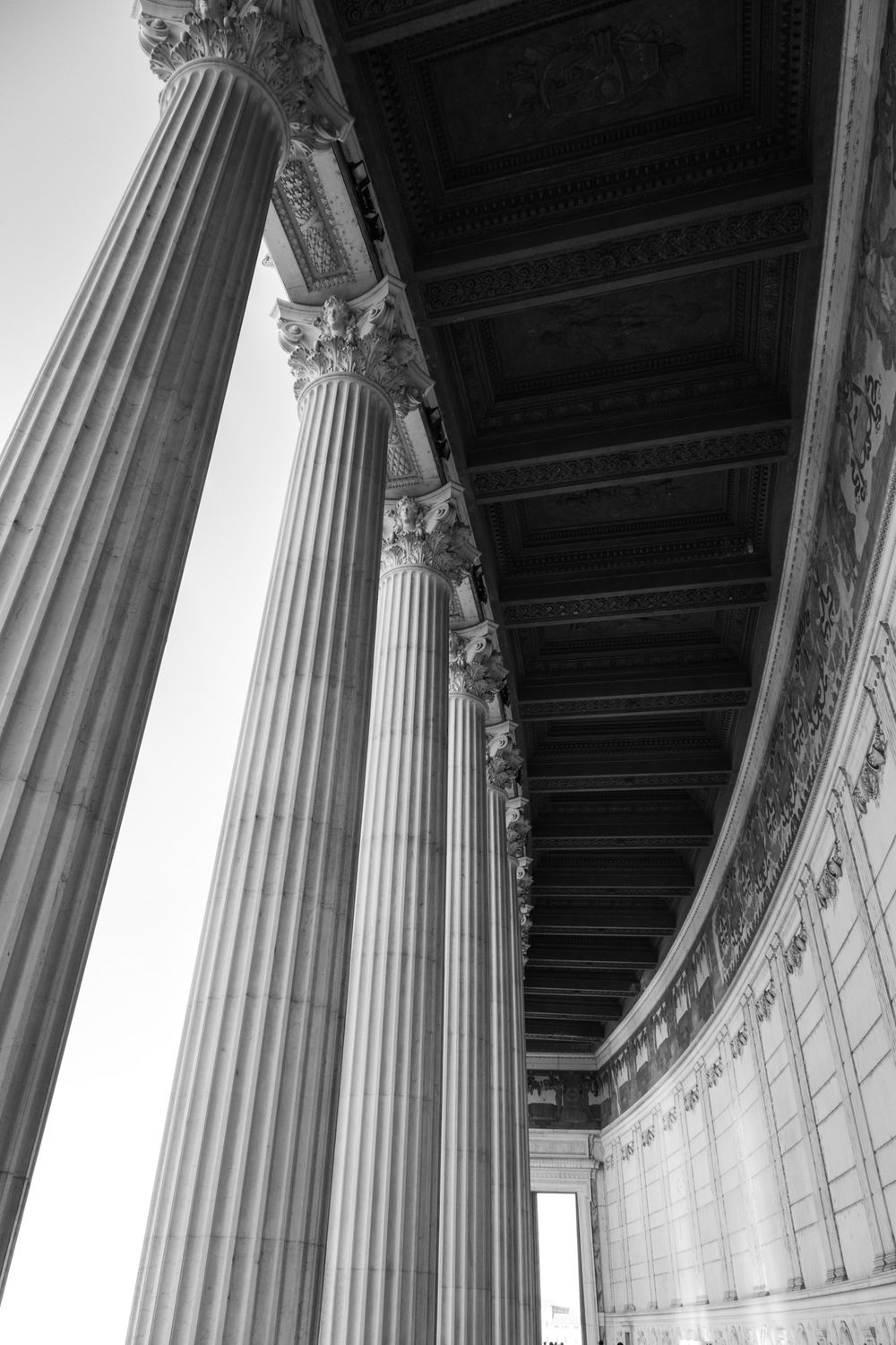 Black and white view of tall fluted Corinthian columns along a curved colonnade with a coffered ceiling and ornate carved frieze.