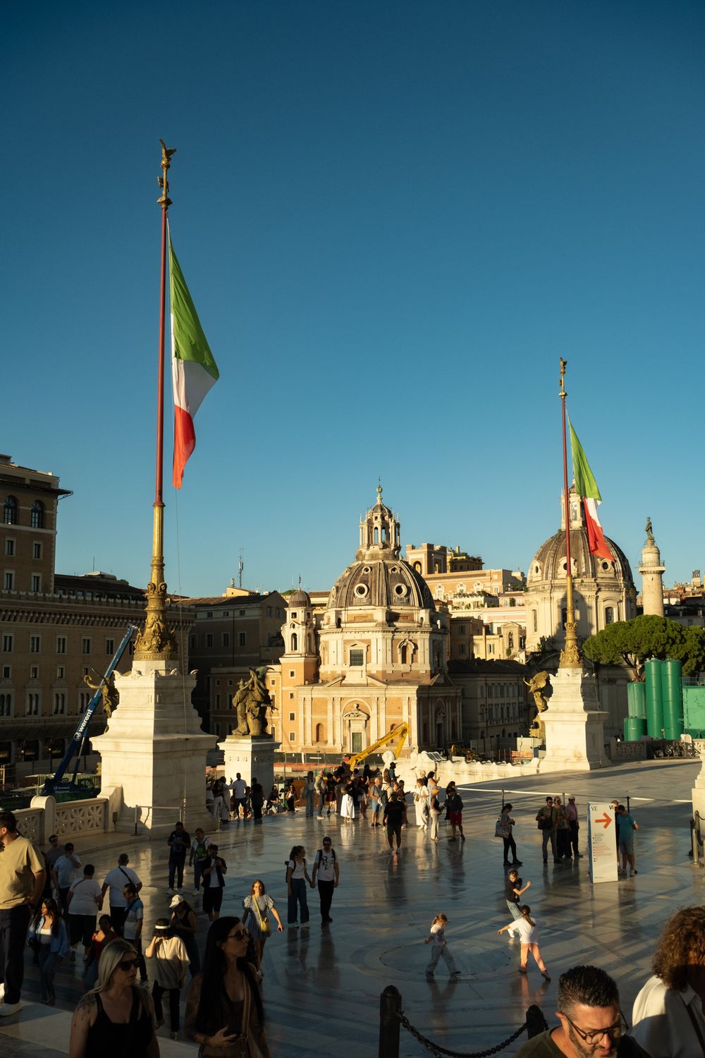 Tourists gather in a sunlit plaza in Rome beneath Italian flags, with baroque domed churches glowing against a clear blue sky.