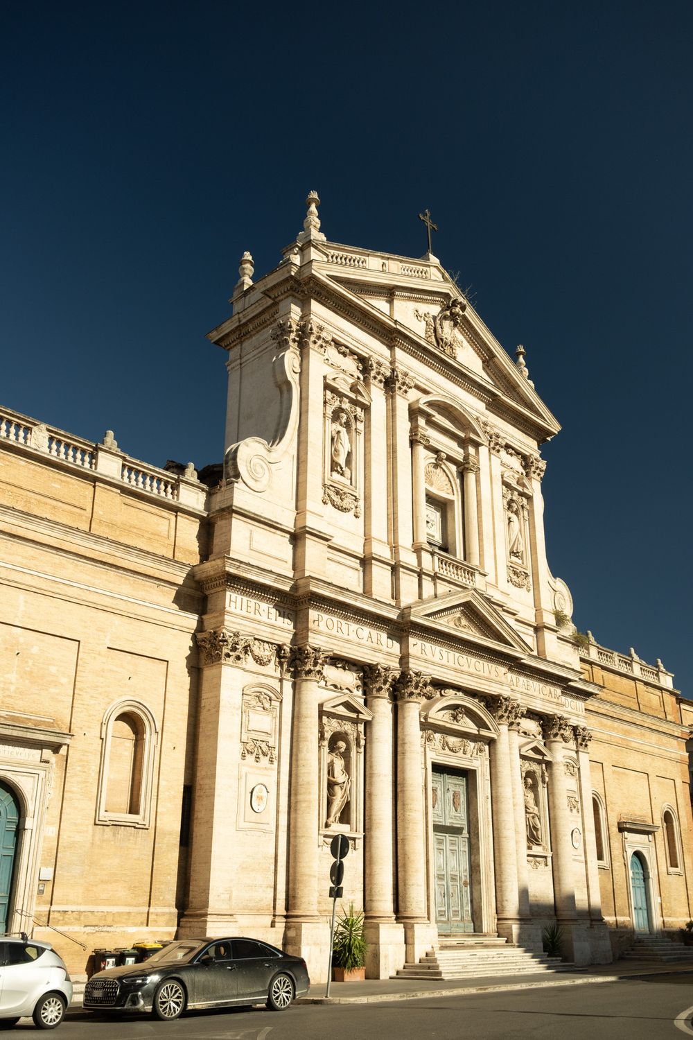 Baroque church facade with ornate columns, statues and a cross on top, glowing in golden light against a deep blue sky in Rome.