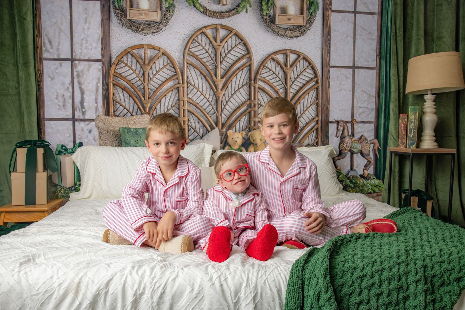 Three children in matching pink striped pajamas sit on a white bed with a green knit blanket in a cozy holiday-themed photo setting.