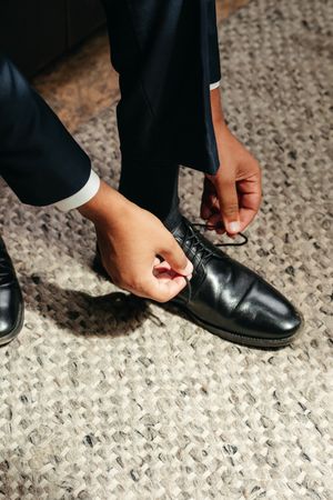 Close-up of a man in a navy suit tying the laces of a polished black leather dress shoe on a woven textured rug.