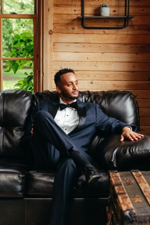 Groom in a navy tuxedo and black bow tie sits on a black leather couch in a rustic wood-paneled room with a vintage trunk.