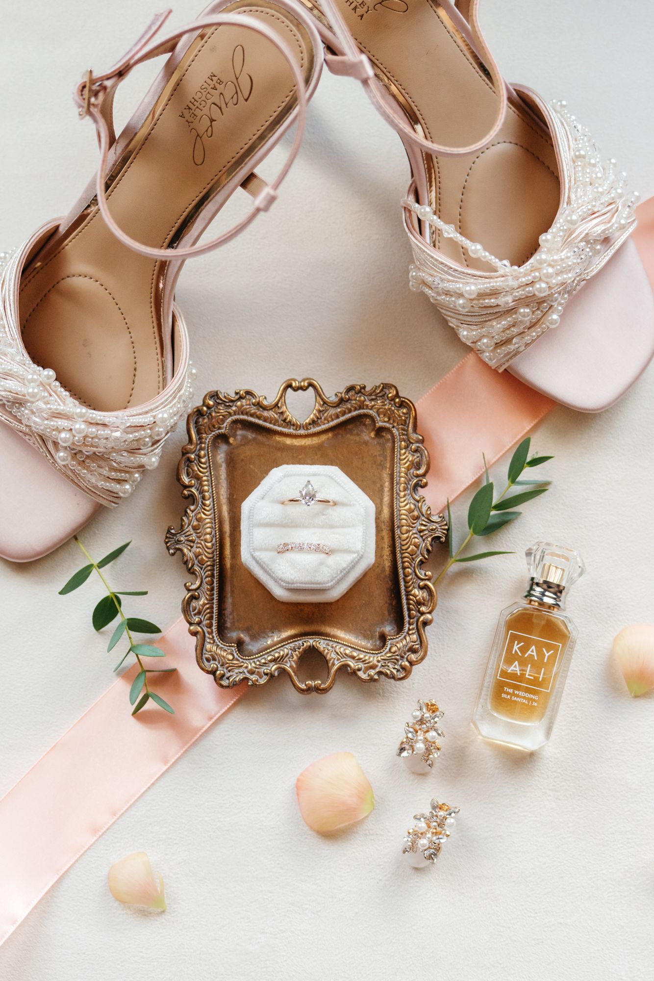 Blush pearl-embellished wedding sandals beside an open ring box on a gold tray, Kay Ali perfume, peach ribbon and rose petals.