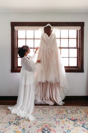 Bride in a sheer white robe adjusts a blush lace wedding gown hanging in a window, above a colorful vintage rug.