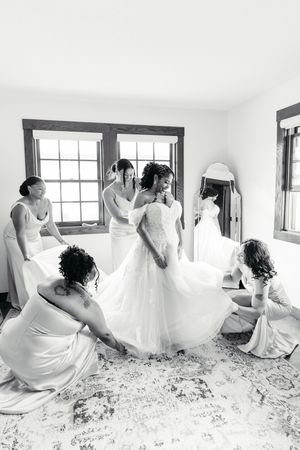 Bridesmaids in satin dresses help a bride in an off-the-shoulder lace gown get ready in a bright, window-lit room in black and white.