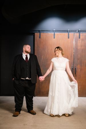 Bride in a lace wedding gown and groom in a dark vest and burgundy tie hold hands and smile at each other before rustic sliding barn doors.