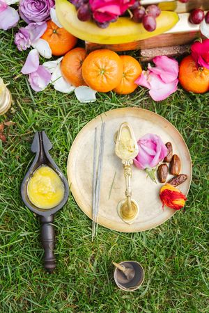 Overhead view of a brass ritual plate with rose, dates and incense sticks, beside a ghee lamp, oranges, grapes and flowers on grass.
