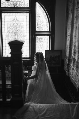 Black and white photo of a bride in a long veil and gown standing by a grand staircase near ornate stained glass windows.