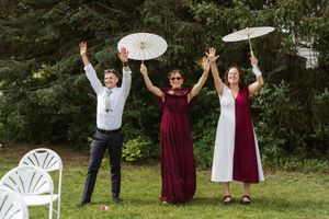 Three people in burgundy and white outfits raise their arms and hold white parasols on a sunny lawn near trees and white folding chairs.