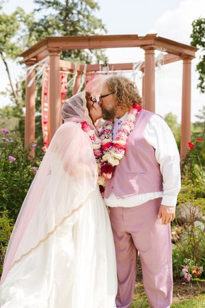 Bride in a white gown and pink veil kisses groom in a pink vest and pants, both wearing floral leis, in front of a wooden garden gazebo.