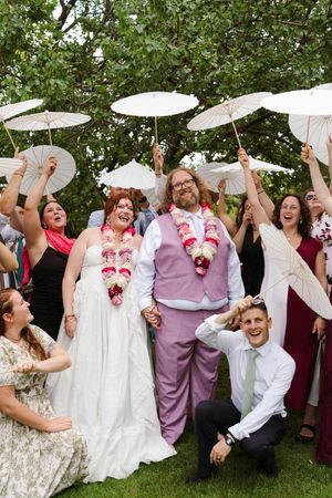 Smiling wedding couple in floral leis holds hands while guests hold white parasols around them on a sunny lawn, one man kneeling in front.