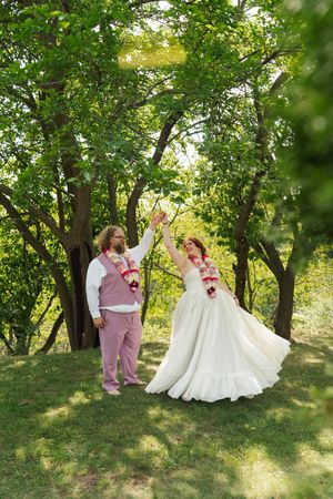 Bride in a flowing white gown and groom in a pink vest and pants dance outdoors on a sunlit lawn under green trees, wearing floral garlands.