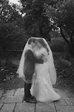 Black and white photo of a bride and groom embracing forehead to forehead under a sheer wedding veil on a stone path in a garden.
