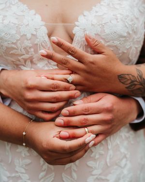 Close-up of two couples' hands with wedding rings resting on a bride's white lace wedding dress, one hand showing a leaf tattoo.