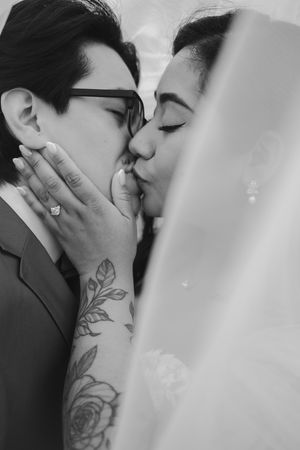 Black and white close-up of a bride in a sheer veil kissing her groom in glasses, his rose-tattooed arm and engagement ring visible.