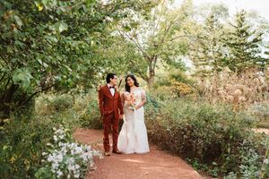 Bride in a white gown holding a bouquet walks hand in hand with a groom in a rust suit along a garden path surrounded by lush greenery.