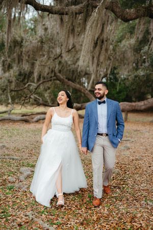 Smiling bride in a white tulle gown holds hands with a groom in a blue blazer and bow tie, walking under moss-draped oak trees.