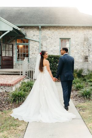 Bride in a flowing white gown and groom in a navy suit hold hands on a walkway outside a rustic stone wedding venue.