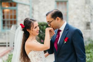 Groom in a navy suit and burgundy tie kisses the hand of his smiling bride, who wears a lace wedding dress, veil and red flower in her hair.