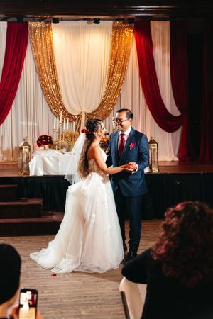 Bride in a white tulle gown and groom in a navy suit share a first dance on a stage with gold sequin and red velvet drapes.