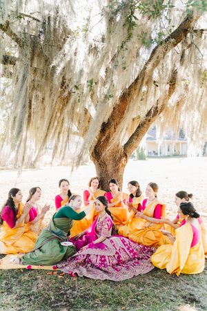 Bride in a magenta embroidered lehenga sits under a Spanish moss-draped oak tree surrounded by women in orange and pink saris.