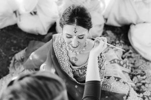 Black and white photo of a bride in traditional Indian attire and jewelry having eye makeup applied by an artist before her wedding.
