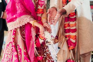 Bride in pink embroidered saree and groom in cream sherwani with floral garlands pour rice during a traditional Indian wedding ceremony.